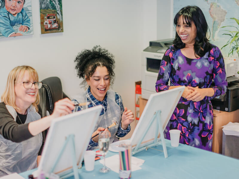 Two women sit at easels, a third woman stands next to them, teaching them to paint